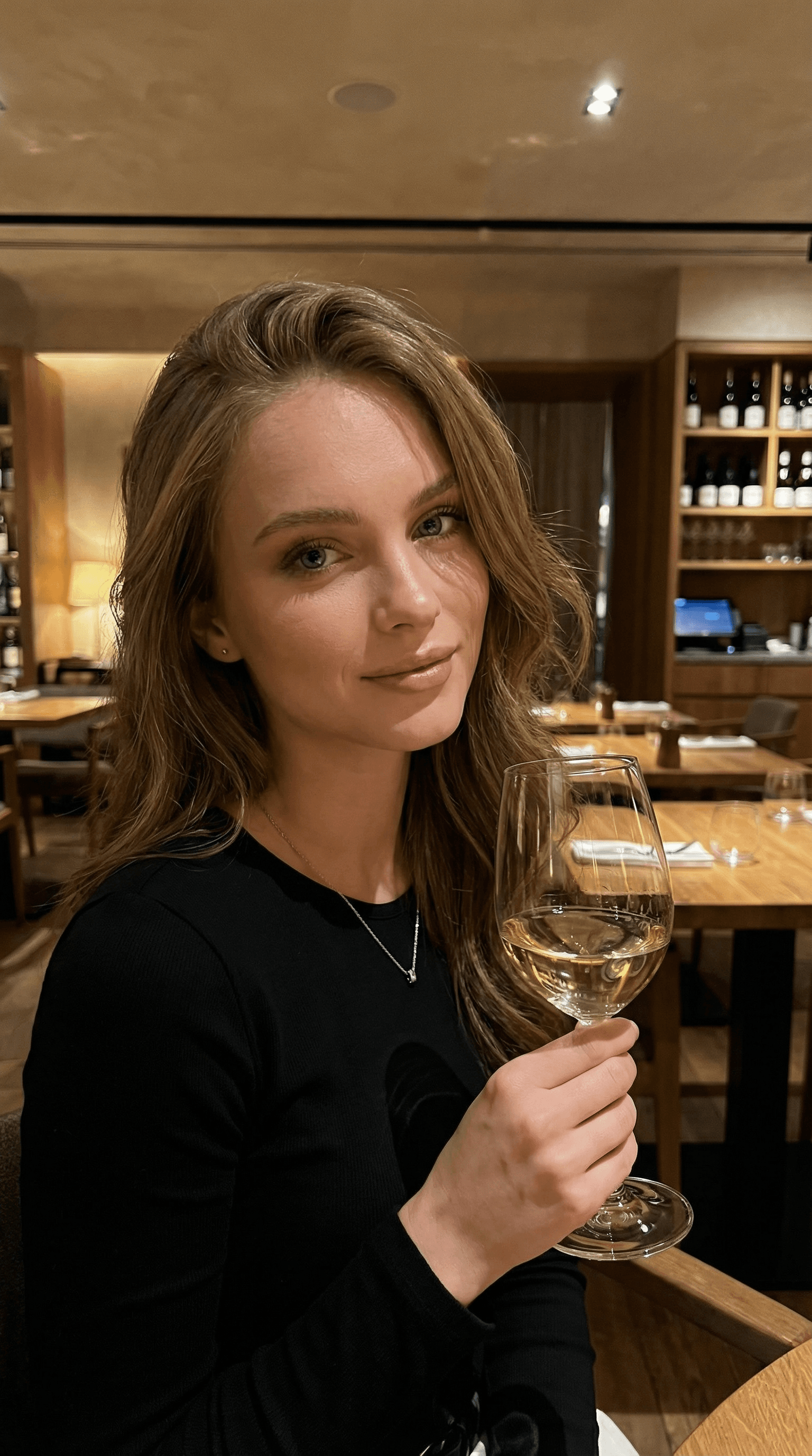 “Replace the person in the image with a hyper-realistic female AI character that fully matches all previously approved parameters and corrections (face, body proportions, skin tone, overall style and realism).
Use the provided reference images to exactly replicate the outfit and accessories, including fabric, color, fit, and details.
Place the character in the restaurant location shown in the reference images, maintaining accurate architecture, furniture, lighting, and atmosphere.

The character is seated on a chair or armchair, slightly posing with a relaxed, elegant posture.
She has a light, natural hairstyle softly styled to the side, with a balanced, natural hair color matching the reference — no over-blonding, no platinum, no artificial highlights.
Hair color should look soft, natural, slightly warm or neutral, not bright blonde.

She is holding a glass of white wine in one hand.
No phone or iPhone in hands.
No tattoos on the body or skin.

The scene should look like a real photograph taken by another person, not a selfie.
Ensure natural perspective, realistic shadows, correct light direction, and seamless integration with the environment.
High facial detail, natural skin texture, realistic depth of field, no AI artifacts, no distortion, no stylization or cartoon look.