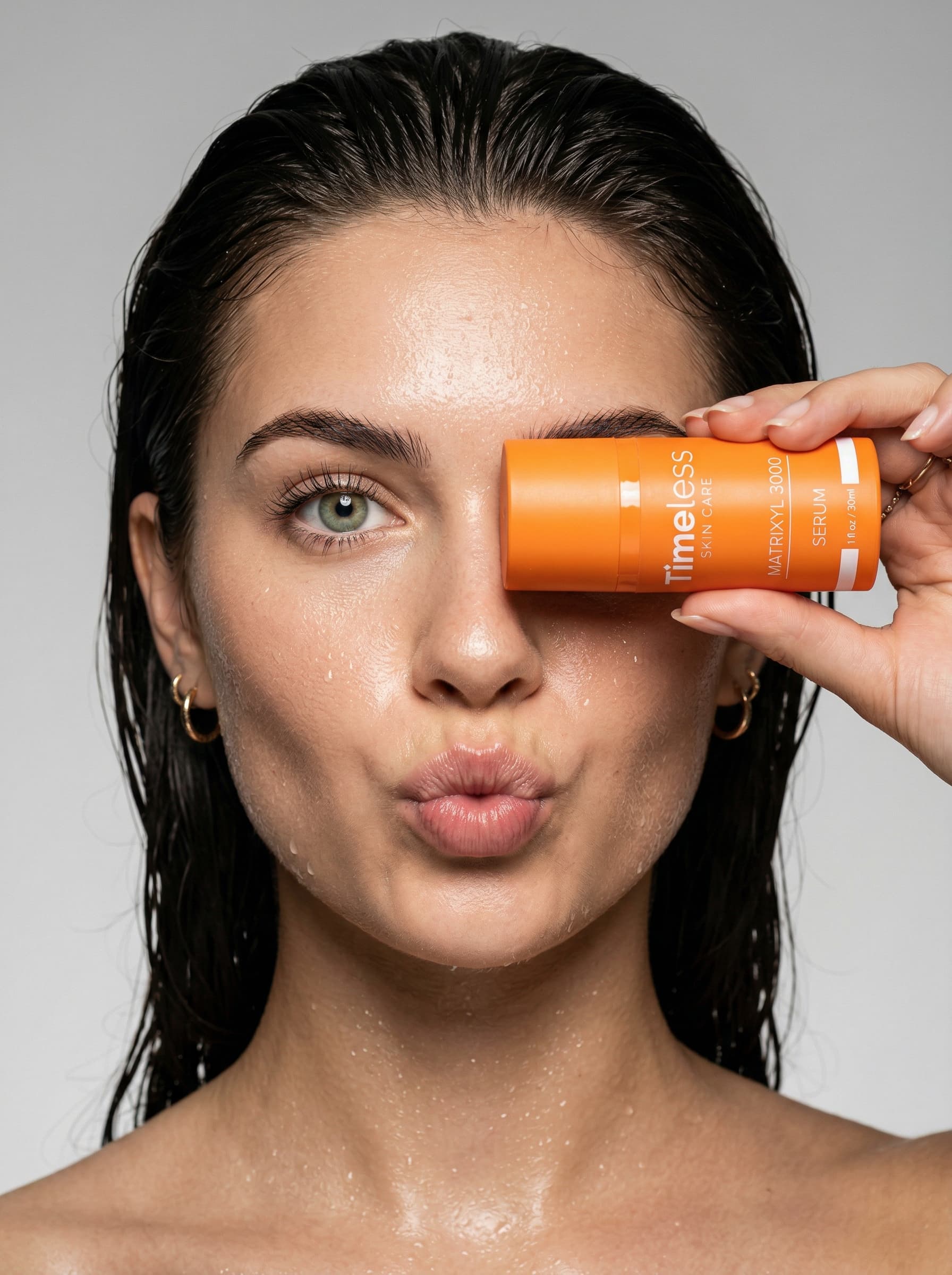 An extreme close-up of a beautiful photograph of a stunning model with light green eyes and sleek, dark, damp hair slicked back. She poses playfully, holding a compact, bright orange, cylindrical bottle of "Timeless Skin Care" (measures 10.8 x 4.6 cm) horizontally in front of her right eye, completely covering it. Shot on a Sony A7R IV with a Sony FE 90mm f/2.8 Macro G OSS lens to capture every detail. Her left eye is open, looking directly at the camera. Her lips are playfully pursed in a "pout." Her skin is radiant thanks to the "glass skin" effect and fine water droplets. Lighting: high-contrast studio setup, softbox light from the front, subtle rim light to highlight the silhouette against a neutral, light gray background. Settings: f/5.6 for sharp focus on both product and face, ISO 100, 1/200 sec. High resolution, 8k, hyper-realistic commercial aesthetics